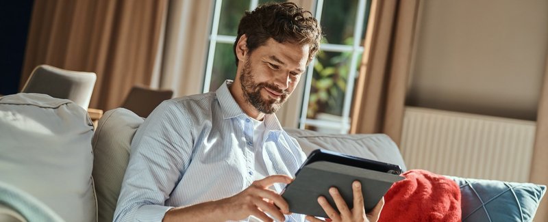 Mężczyzna ubrany w koszulę, siedzący na kanapie w salonie, trzyma w rękach tablet na którym pracuje