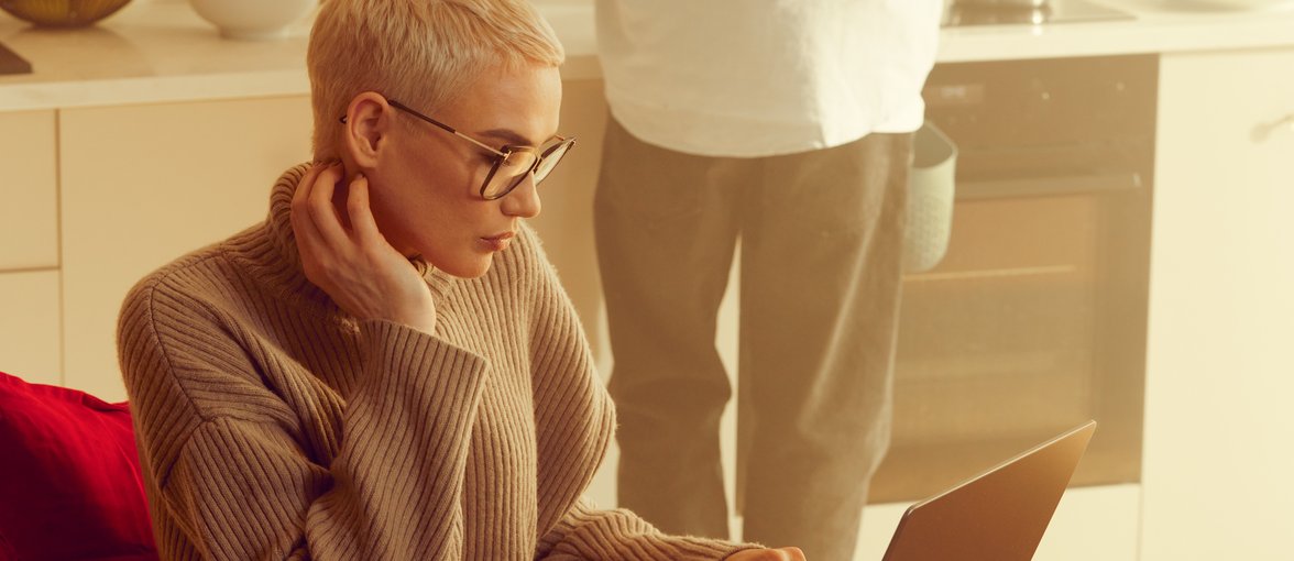 Blond kobieta o krótkich włosach siedzi przy stole i pracuje na komputerze, w tle widać mężczyznę który gotuje na sprzęcie Samsunga