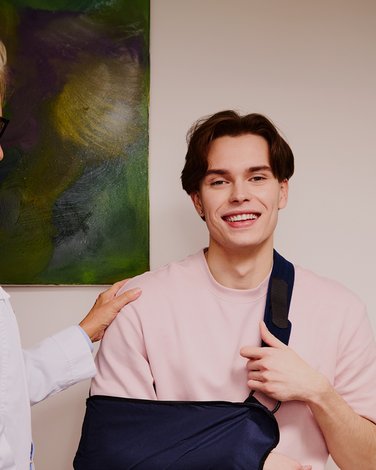 Mężczyzna, z usztywnioną ręką uśmiecha się do kamery, obok niego stoi lekarz.