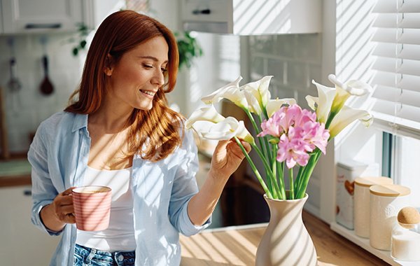 Kobieta w kuchni trzyma kubek i patrzy na świeże kwiaty w wazonie.