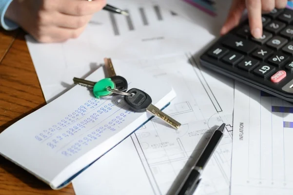 Notes z kwotami, klucze, kalkulator i długopis znajdujące się na biurku. W tle dłonie: osoba podpisująca dokumenty i dokonująca kalkulacji. Blog