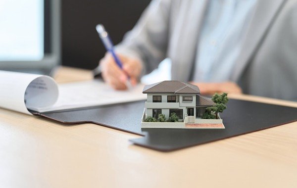 Model domu na biurku, w tle osoba podpisuje dokumenty. Blog.