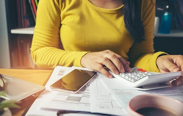 Kobieta w żółtym swetrze siedzi przy biurku, używa kalkulatora i analizuje dokumenty finansowe. Na stole leżą telefon, okulary, laptop i filiżanka kawy. Blog.