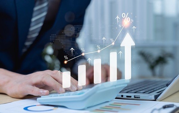 Osoba korzysta z kalkulatora przy laptopie, na tle wykres rosnących odsetek i symbol procentu. Blog.