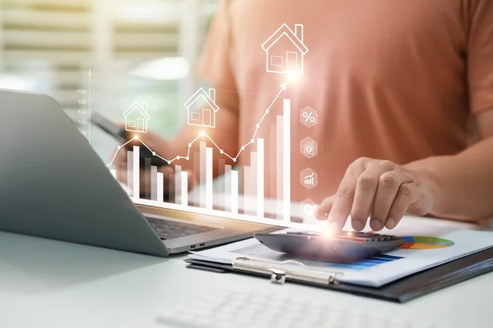 Osoba wykonująca obliczenia przy pomocy kalkulatora i laptopa. Między osobą a laptopem pojawia się wizualizacja wykresu z symbolami hipotecznymi. Blog