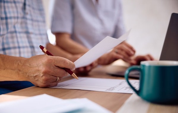 Osoba trzyma dokument i długopis przy biurku, w tle laptop i kubek. Blog.