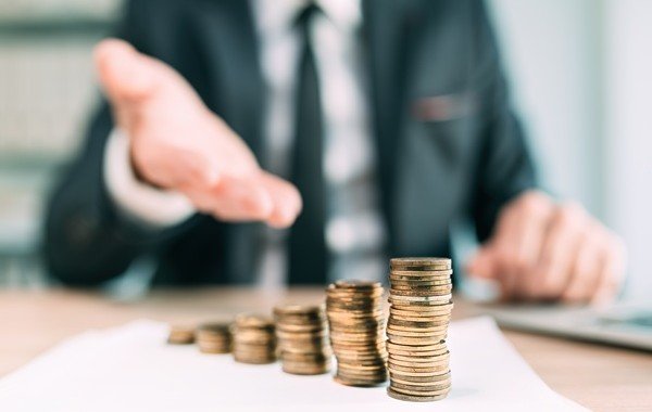Stosy monet na biurku, w tle osoba w garniturze wyciąga rękę – symbol oszczędności i ulg podatkowych przy kredycie hipotecznym. Blog.