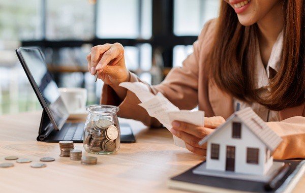 Kobieta odkłada monety do słoika, obok rachunki i model domu na biurku. Blog.