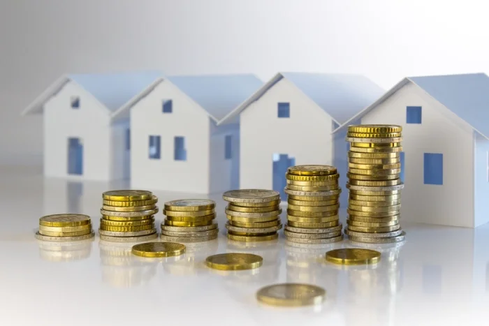 Stosy monet znajdujące się obok małych makiet wizualizujących domy jednorodzinne, symbolizujące oszczędności i inwestycje w nieruchomości. Blog